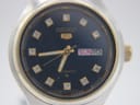 Close-up front view of the Seiko 5 black dial with gold-tone hands, stone markers, and day-date window.