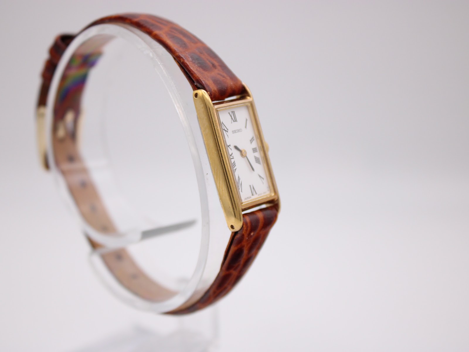 Three-quarter view of the Seiko watch on its clear display stand, showcasing the gold case and brown leather strap.