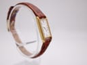Three-quarter view of the Seiko watch on its clear display stand, showcasing the gold case and brown leather strap.