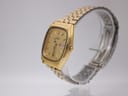 Side-angle view of the gold Seiko watch and integrated steel bracelet on a clear display stand, showing the crown.