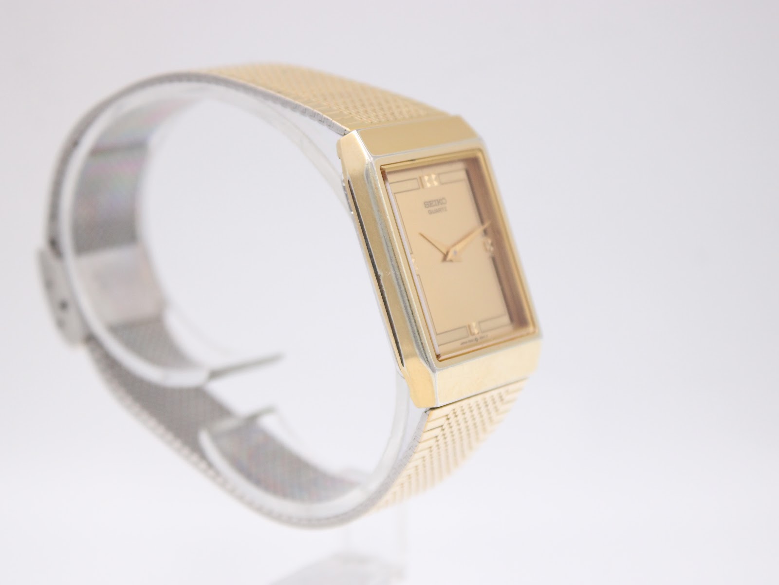 Angled side view of the gold tone Seiko watch on a clear stand, highlighting its slim rectangular case.