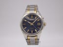 Front view of the Seiko 5M62-0D50 watch, showing the navy dial, gold-tone indices, and two-tone steel bracelet on a stand.