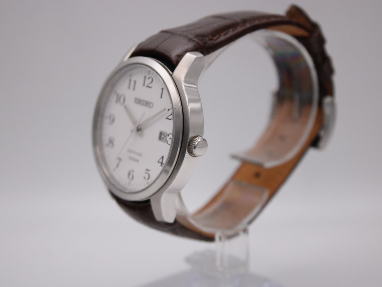 Side view of the Seiko watch, showing the polished stainless steel case, crown, and brown leather strap on a clear stand.
