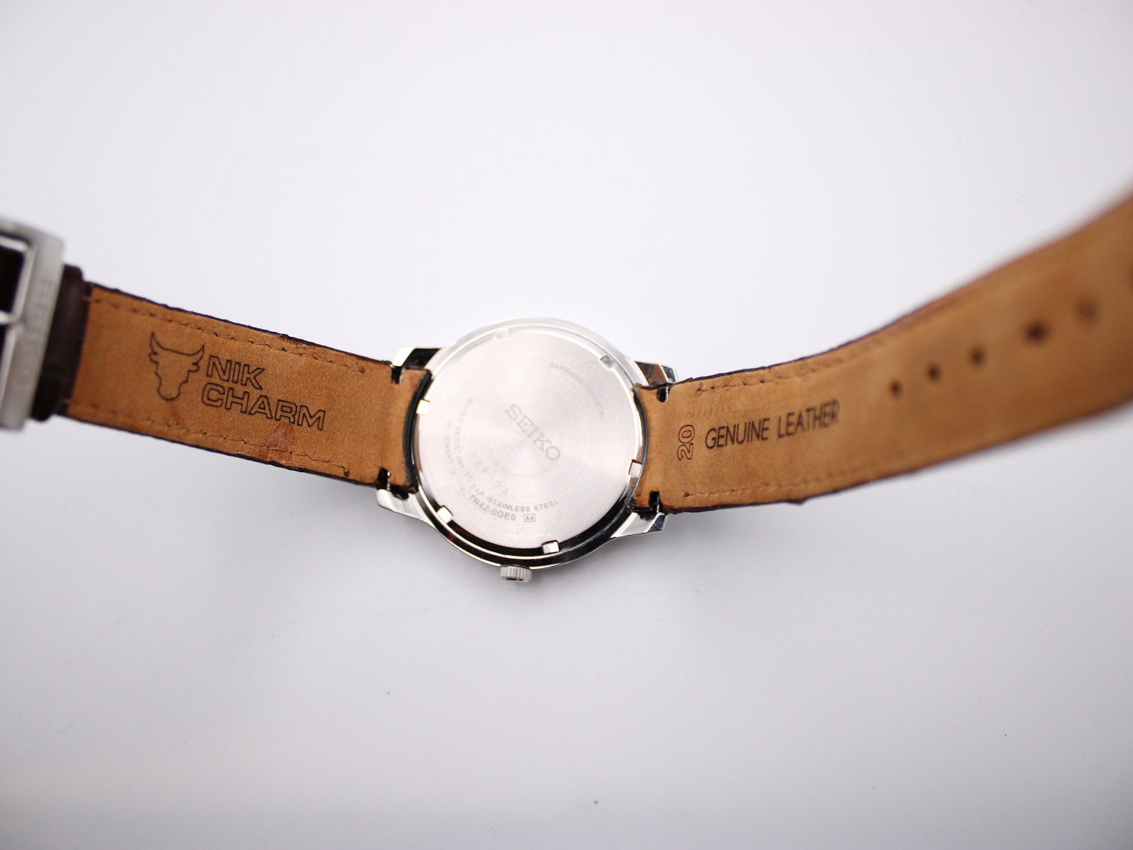 Interior view of the brown leather strap, showing 'NIK CHARM' and 'GENUINE LEATHER 20' markings, with the case back in the centre.
