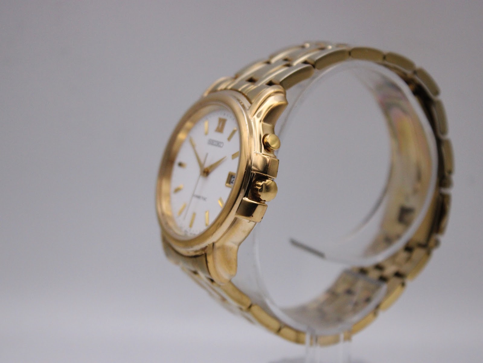 Angled side view of the gold-tone Seiko kinetic watch on a clear display stand, showing the case profile and crown.