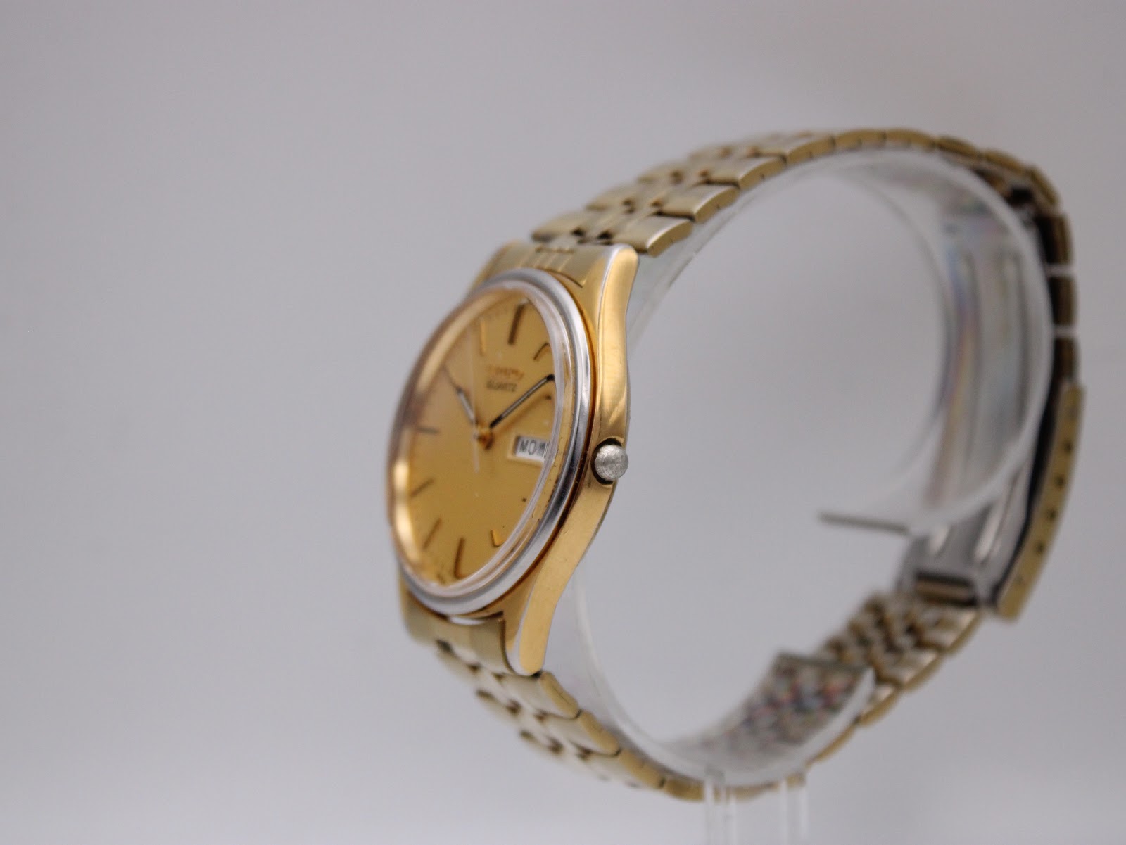 Side profile of the Seiko gold-tone watch on a clear display stand, highlighting the case shape and crown.