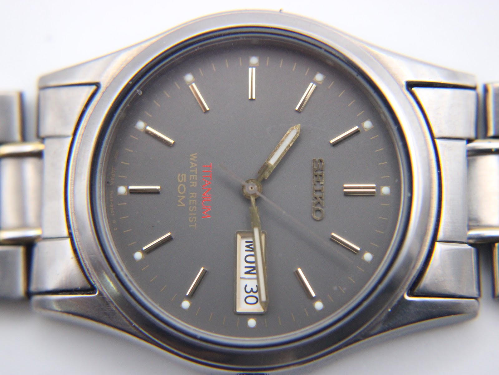 Macro shot of the Seiko watch's dark grey dial, showing gold-tone baton indices and 'Titanium' text.