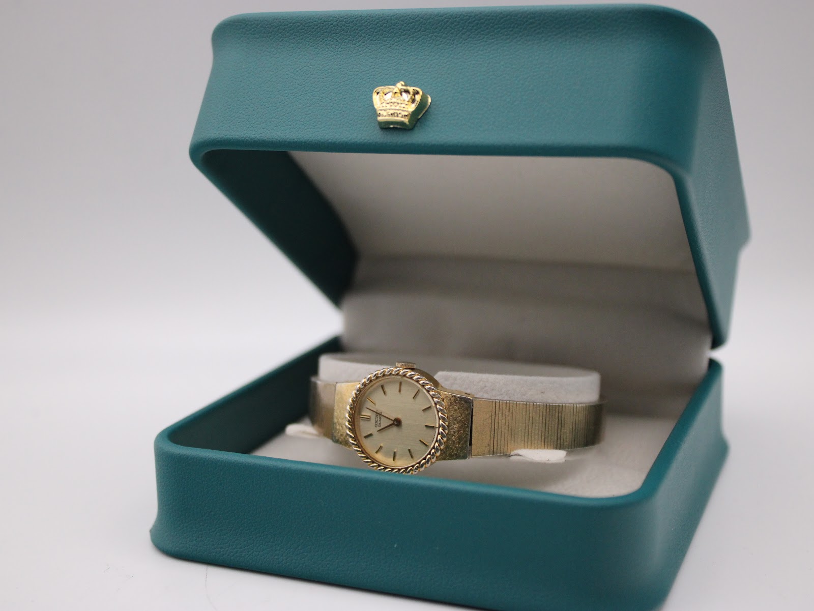 The elegant gold-tone Seiko watch presented inside a teal coloured jewellery box with a distinctive gold crown emblem.