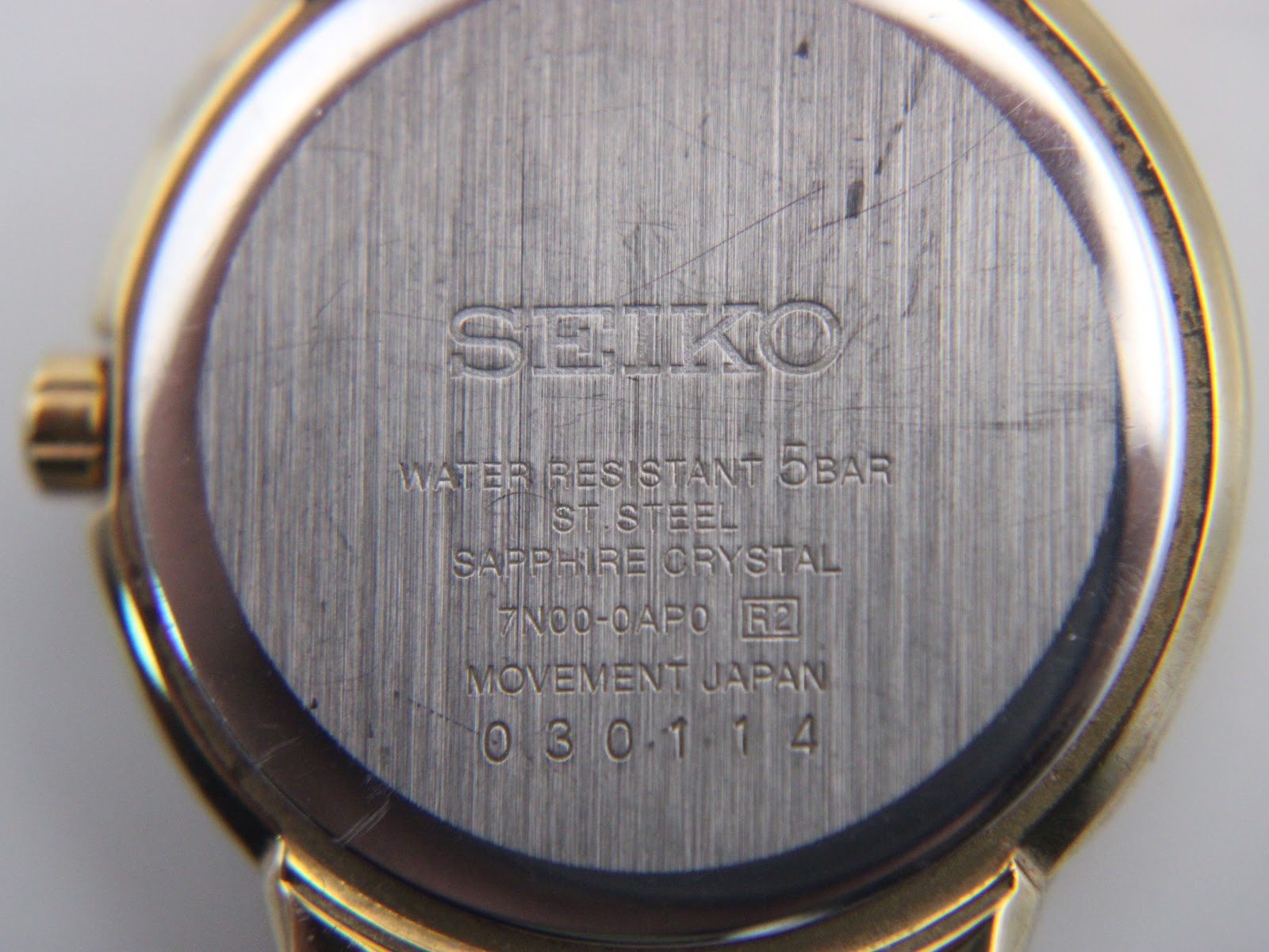 Close-up of the brushed stainless steel case back of the Seiko watch, showing model number, serial, and "sapphire crystal" engraving.