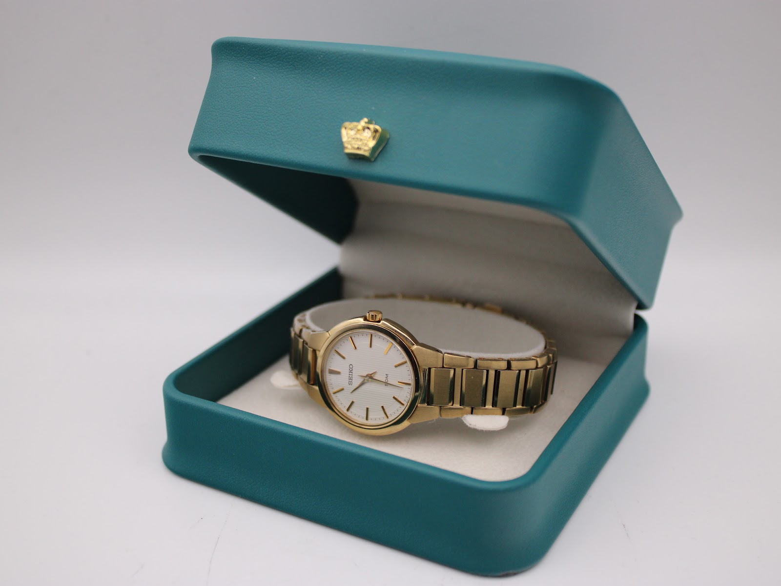 The gold tone Seiko watch displayed inside a teal coloured gift box with a small golden crown decoration on the lid.