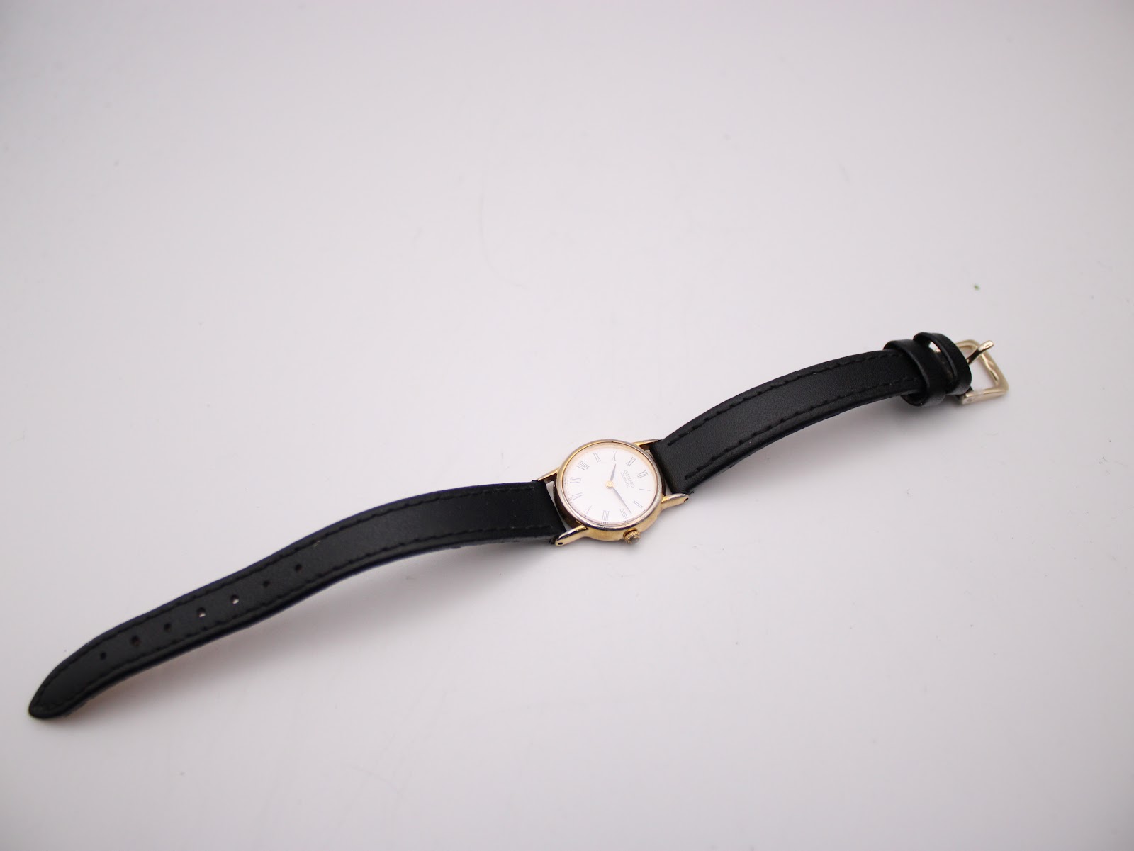 Overhead view of the small gold tone Seiko watch with its black leather strap laid flat on a white surface.