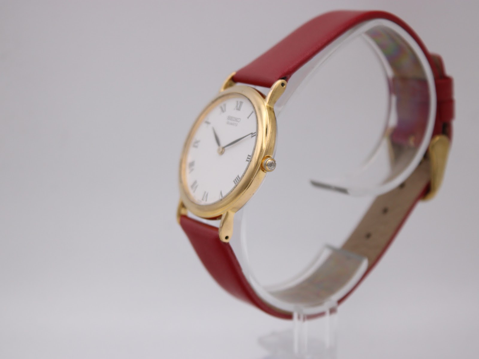 Three-quarter side view of the Seiko watch, showing the profile of the gold-tone case, the crown, and the texture of the burgundy strap on a clear stand.