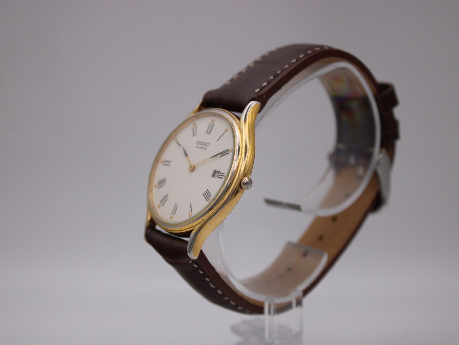 Angled view of the Seiko watch, showing the gold tone case, white dial, and brown leather strap on a watch stand.