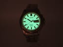 Front view of Seiko Kinetic watch dial glowing in the dark, showing bright green lume on hands and baton markers.