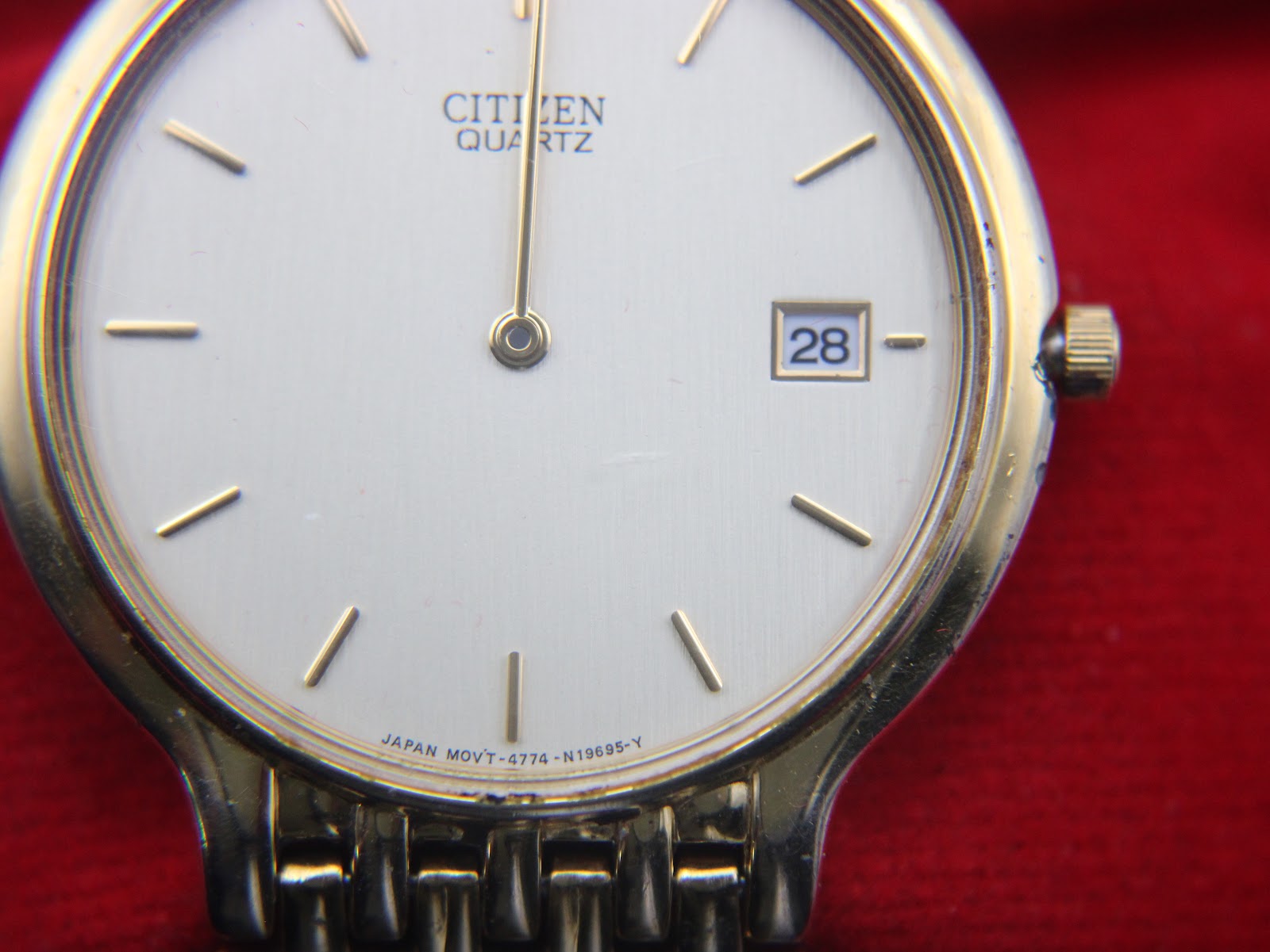 Close-up of the cream dial of the Citizen watch against a red background, showing "Citizen Quartz" text, baton indices, date window, and dial codes.