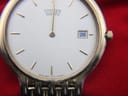 Close-up of the cream dial of the Citizen watch against a red background, showing "Citizen Quartz" text, baton indices, date window, and dial codes.