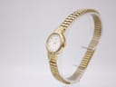 Angled right-side view of the gold tone Citizen watch showing the round case, crown, and expansion bracelet on a display stand.