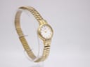 Angled left-side view of the gold tone Citizen watch with the white dial and expansion bracelet on a clear display stand.