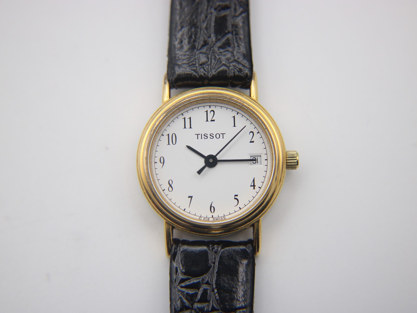 Overhead front view of the Tissot C 212 watch lying flat, displaying the white dial, gold-tone case, and black crocodile-grain leather strap.