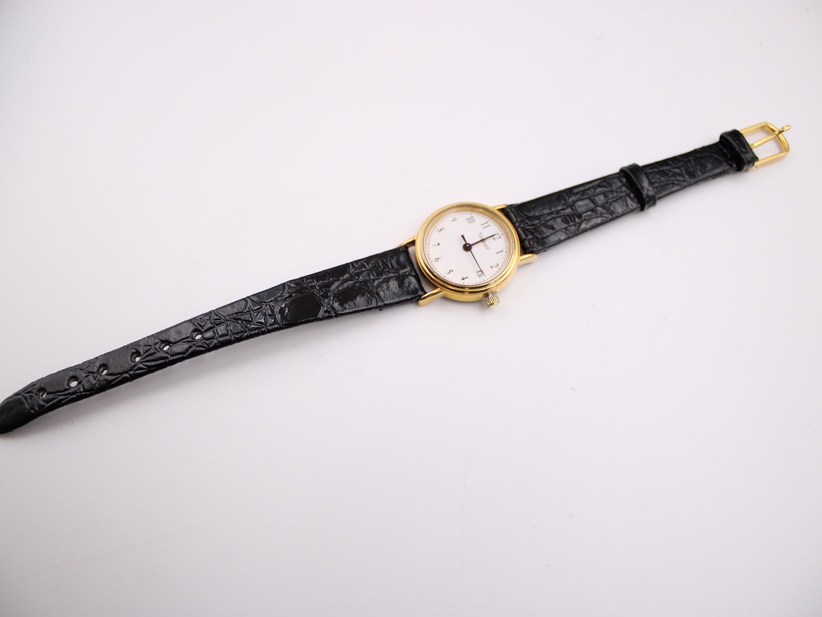 Full view of the Tissot C 212 watch lying flat, showing the black leather strap with its gold-tone pin buckle.