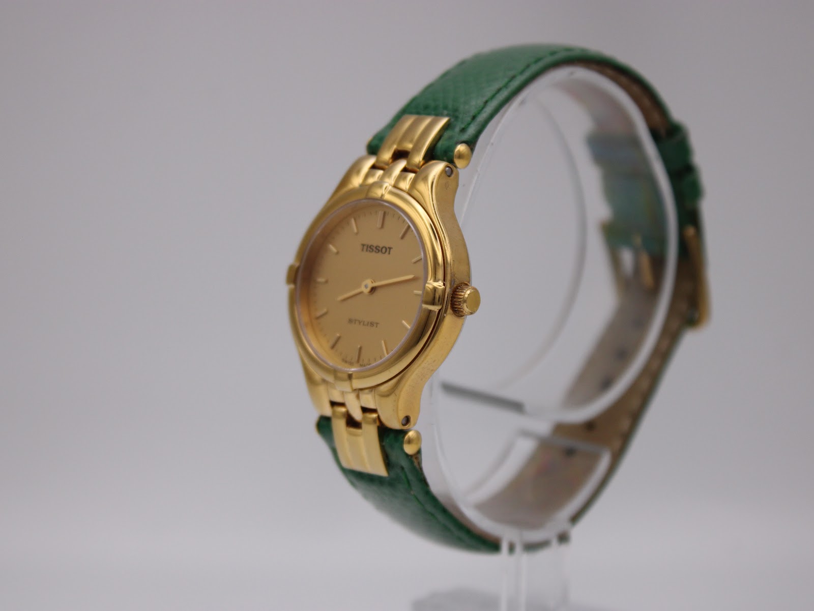 Angled view of the Tissot Stylist watch on a display stand, highlighting the curved gold-tone case and green leather strap.