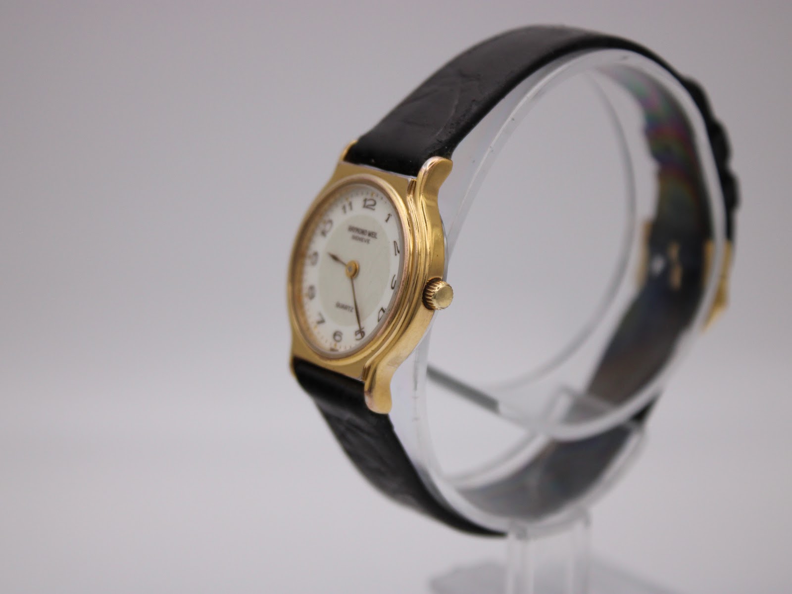Angled side view of the Raymond Weil 5334 watch on a clear stand, showing gold case and black strap.