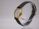 Angled side view of the Raymond Weil 5334 watch on a clear stand, showing gold case and black strap.