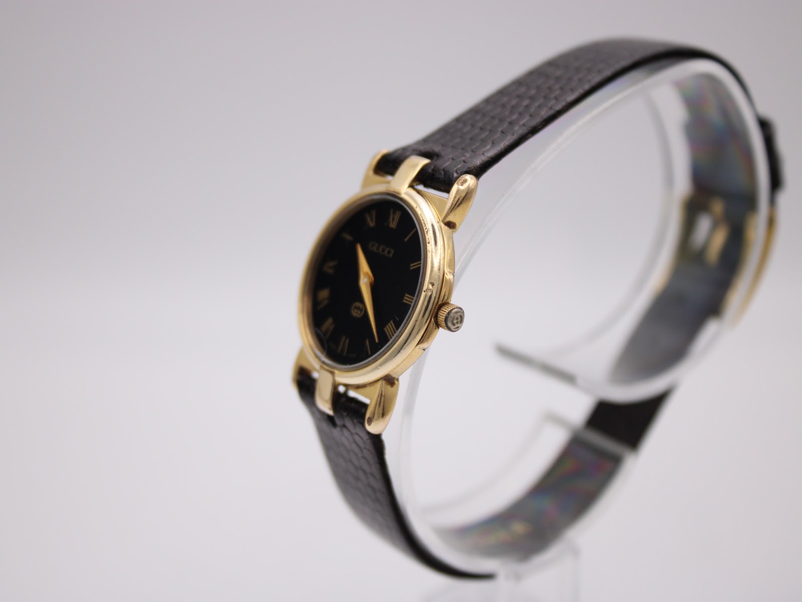 Angled view of the gold-tone Gucci 3400 L watch on a clear display stand, showing the case and black strap.