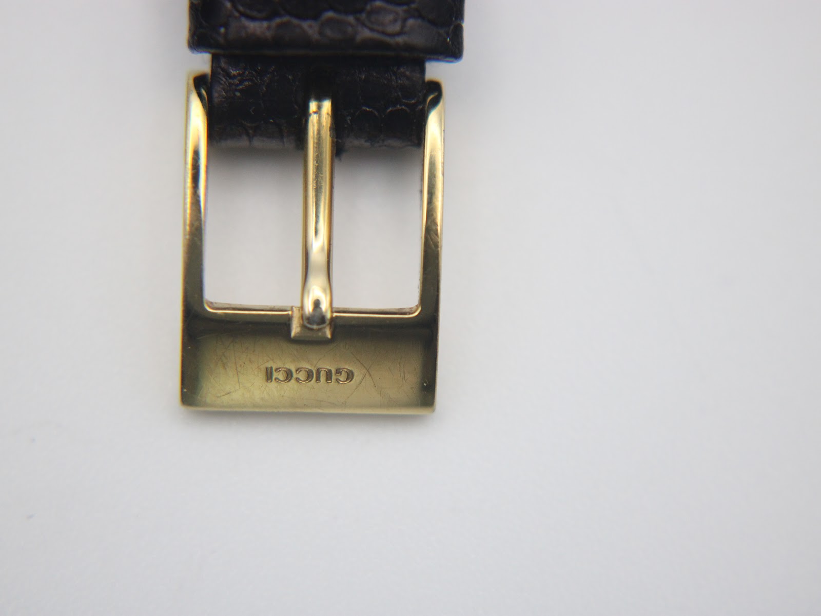 Close-up of the gold-tone buckle on the black leather strap, with "GUCCI" engraved.
