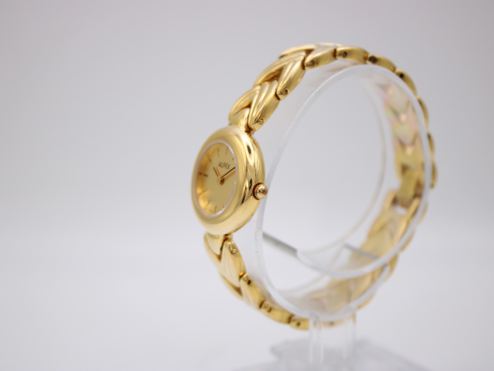 Right side angle view of the Alfex gold watch, showing the round case and crown on the display stand.