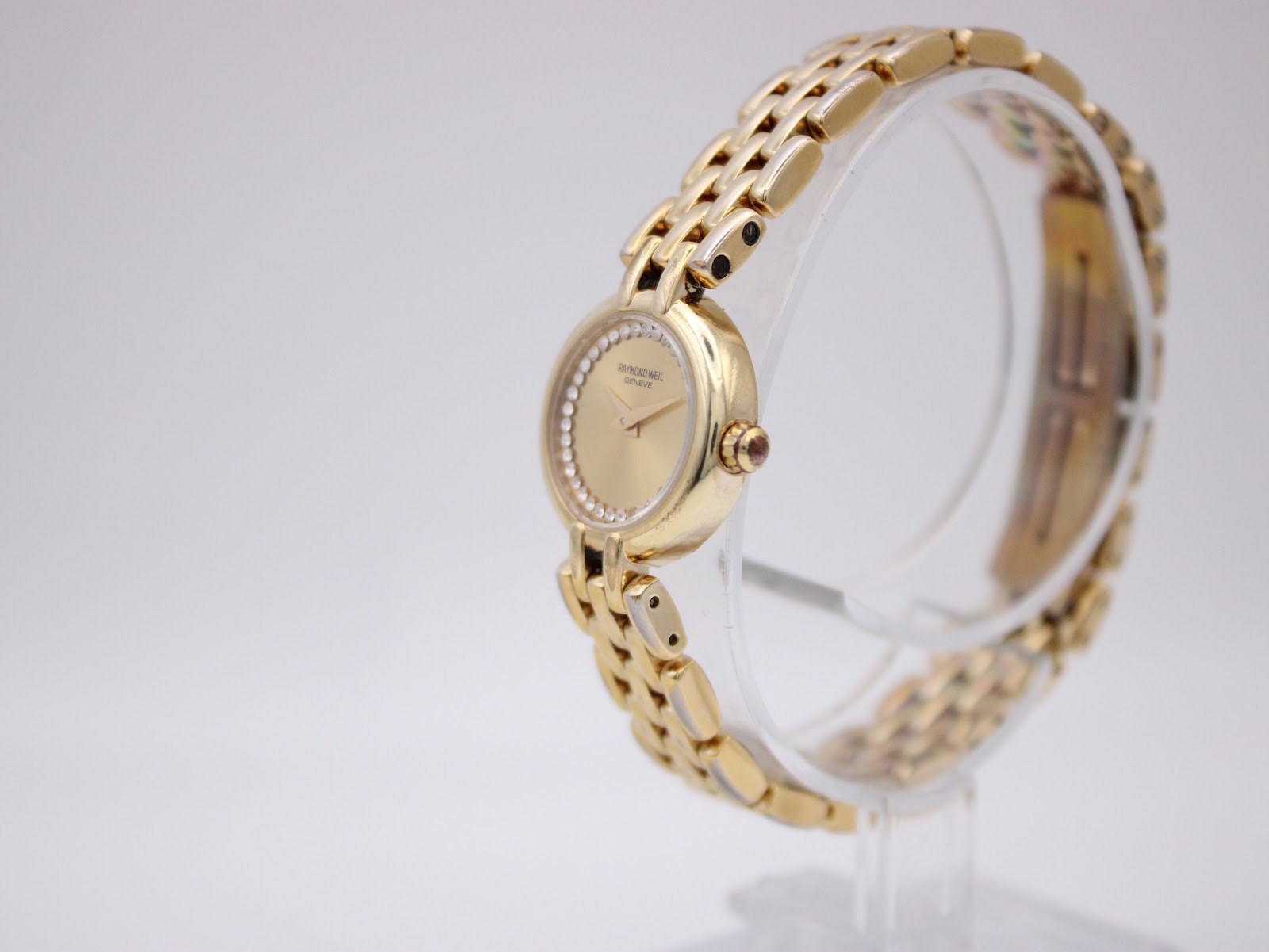 Angled side view of the Raymond Weil watch on a display stand, highlighting the curve of the case and bracelet, with light reflecting off the gold finish.