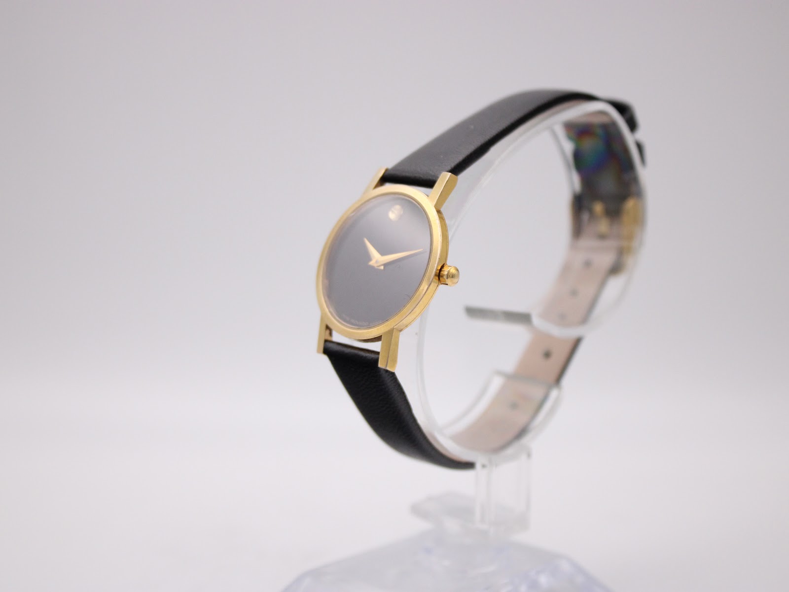Angled view of the Movado watch on a stand, showing the polished gold-tone case and the black leather strap.