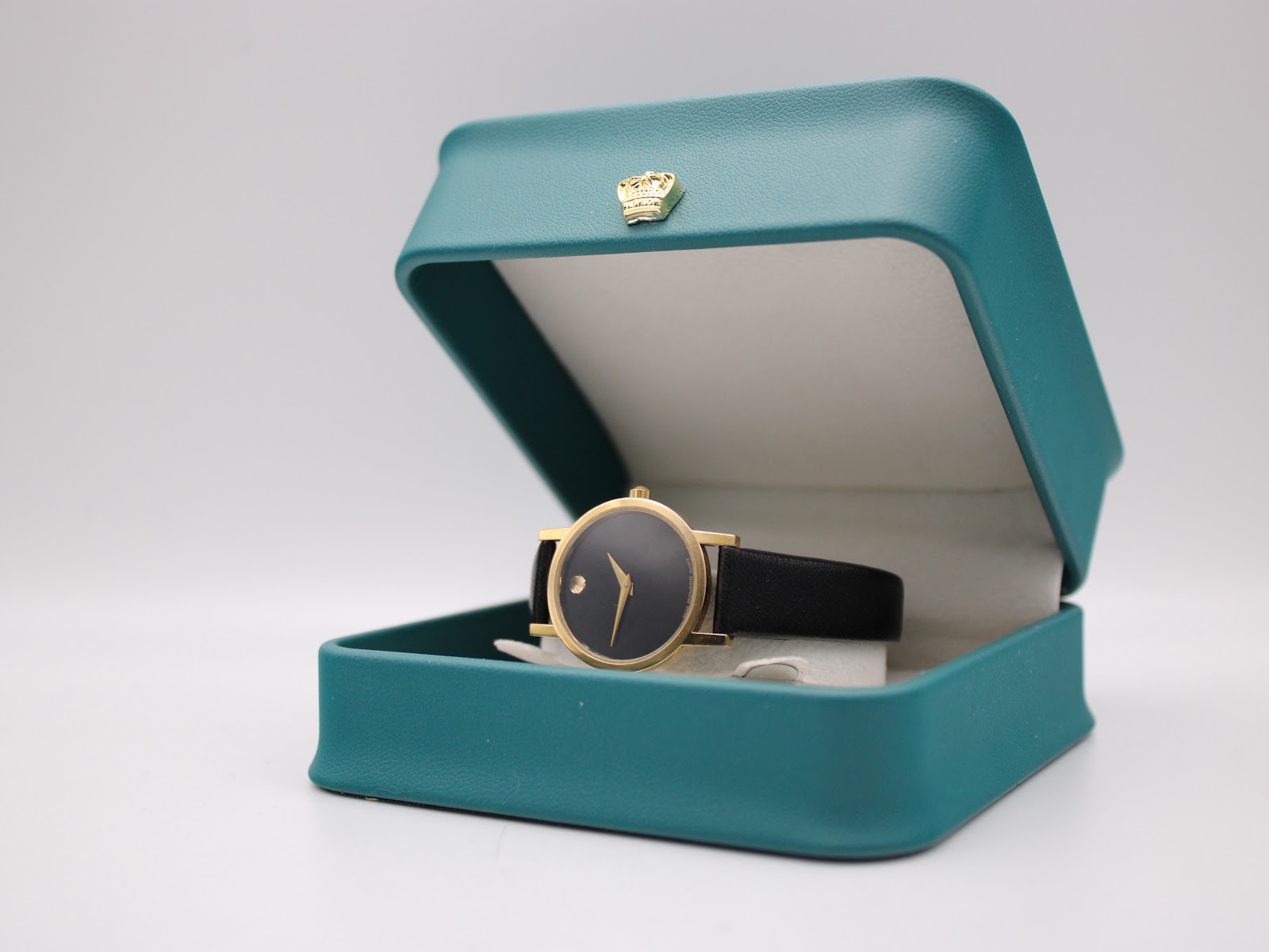 The Movado watch displayed open in a teal presentation box, showing the watch's face and strap.