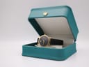 The Movado watch displayed open in a teal presentation box, showing the watch's face and strap.