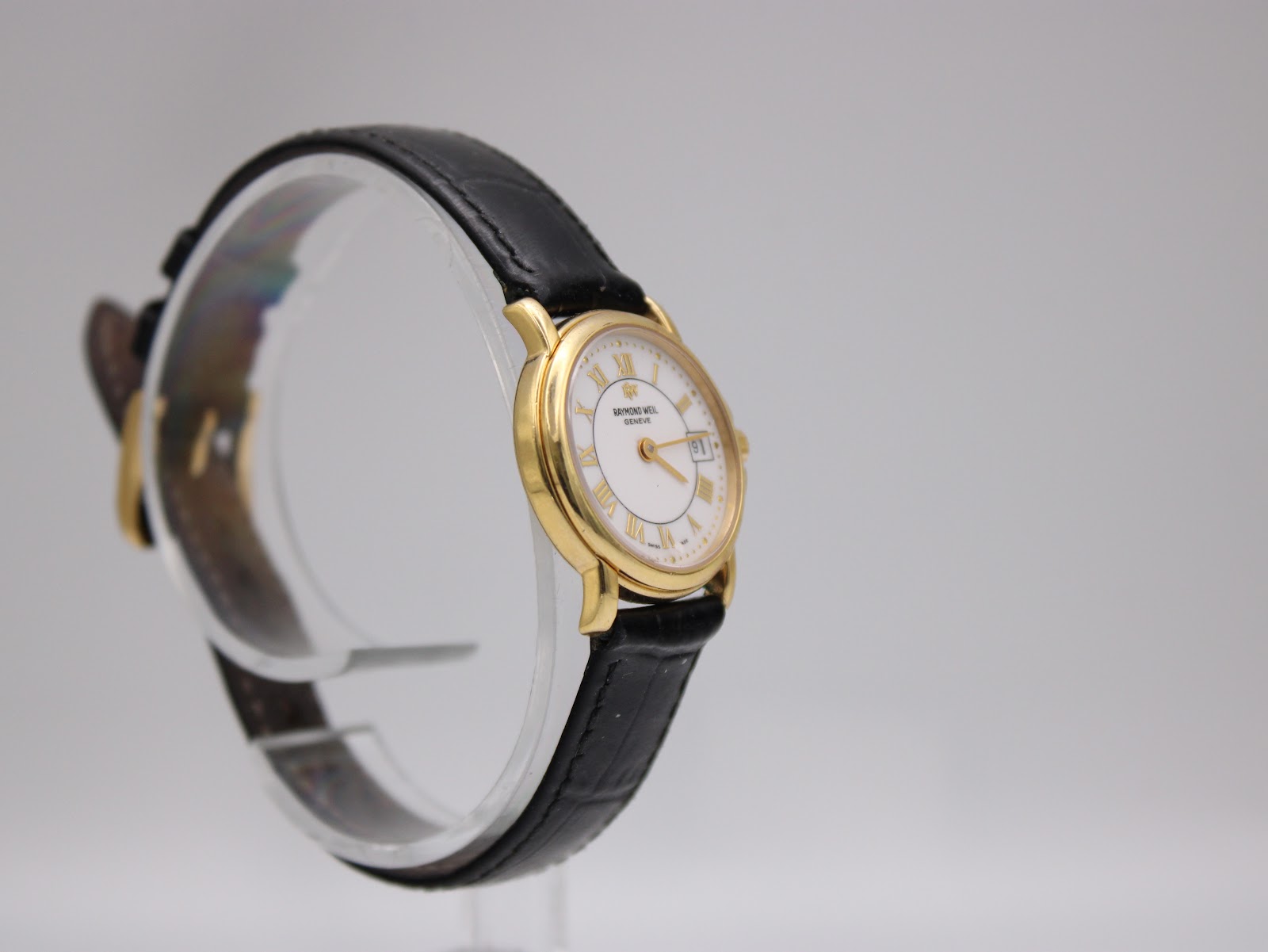 Side view of the Raymond Weil 9972 watch on a clear stand, highlighting the slim profile of the gold plated case and the texture of the black leather strap.