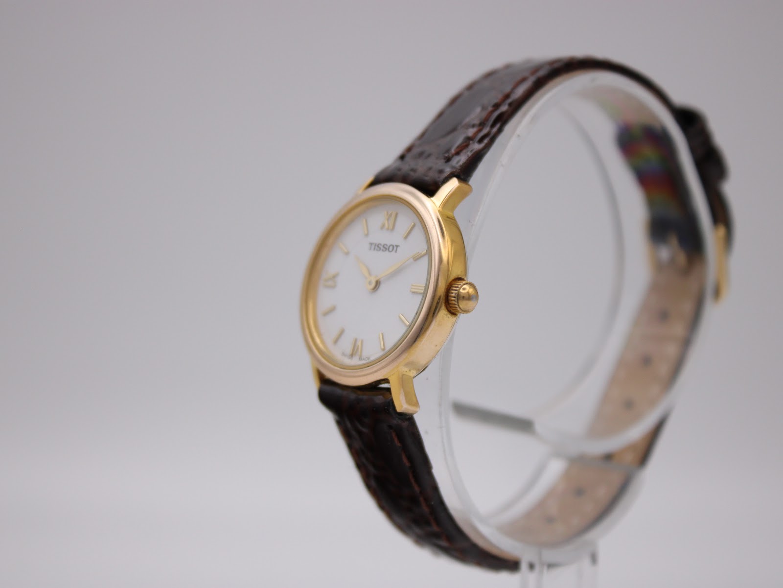 Angled side view of the Tissot C225K watch on a clear stand, highlighting the gold-plated case and crown.
