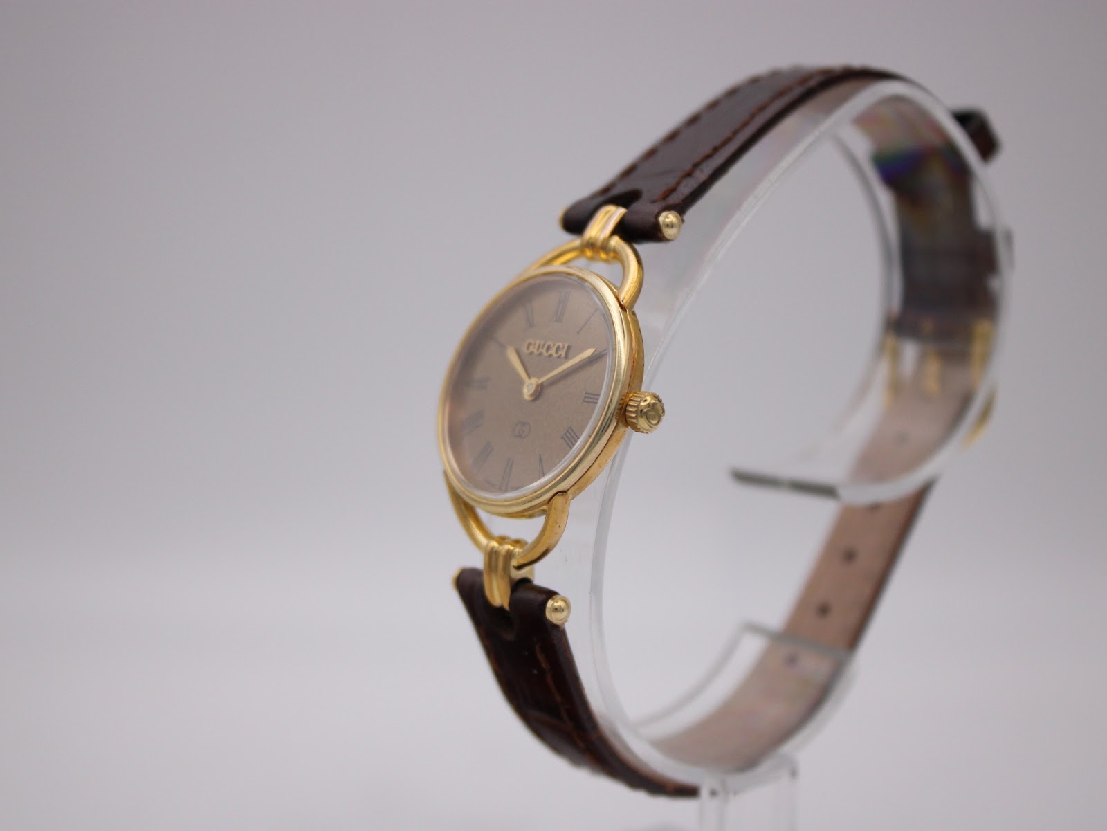Angled side view of the Gucci 6000L watch on a clear stand, highlighting the gold-tone case profile and leather strap.