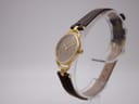 Angled side view of the Gucci 6000L watch on a clear stand, highlighting the gold-tone case profile and leather strap.
