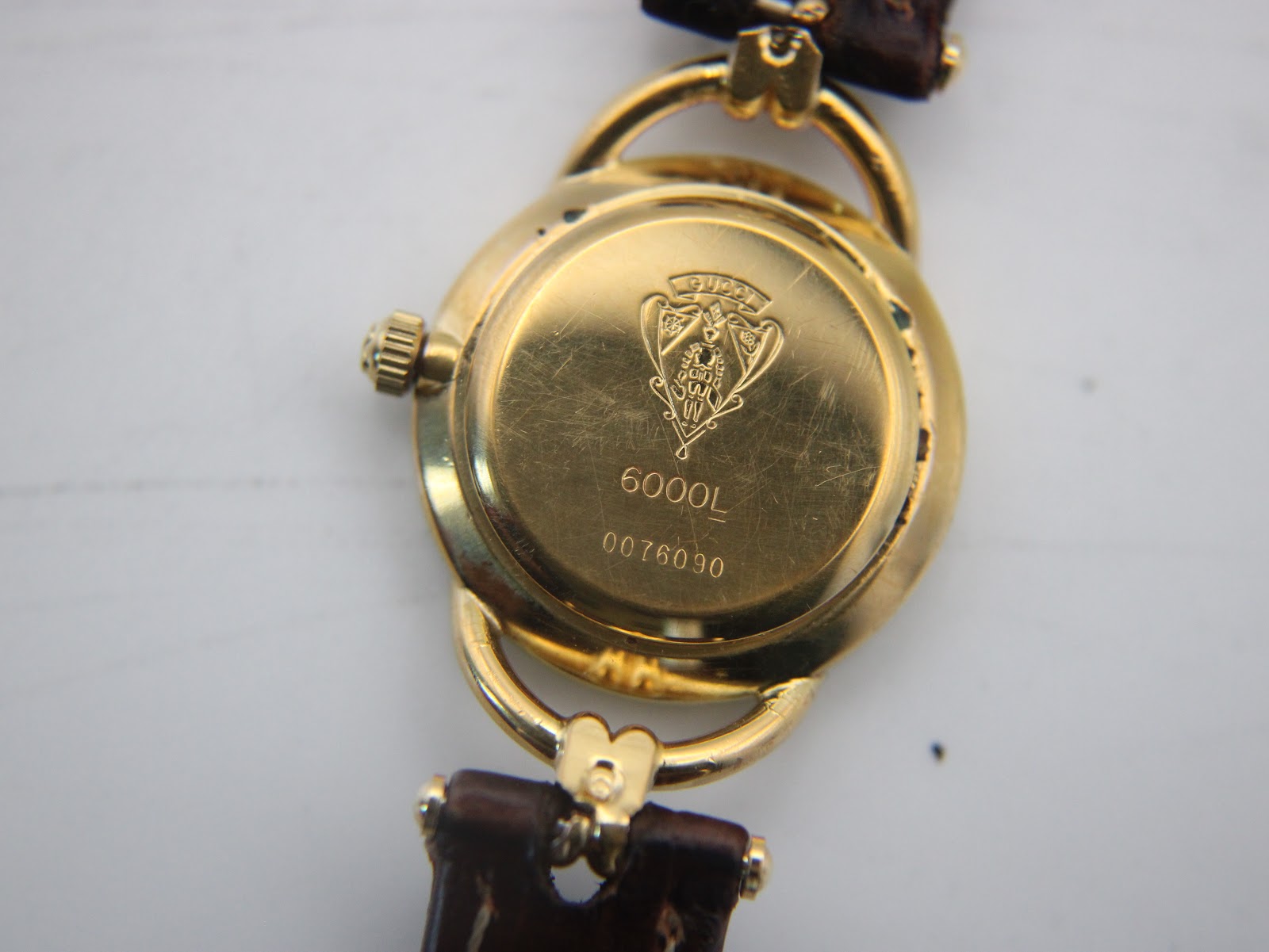 Close-up of the gold-tone case back, engraved with the Gucci crest, model "6000L", and serial number "0076090", showing surface scratches.