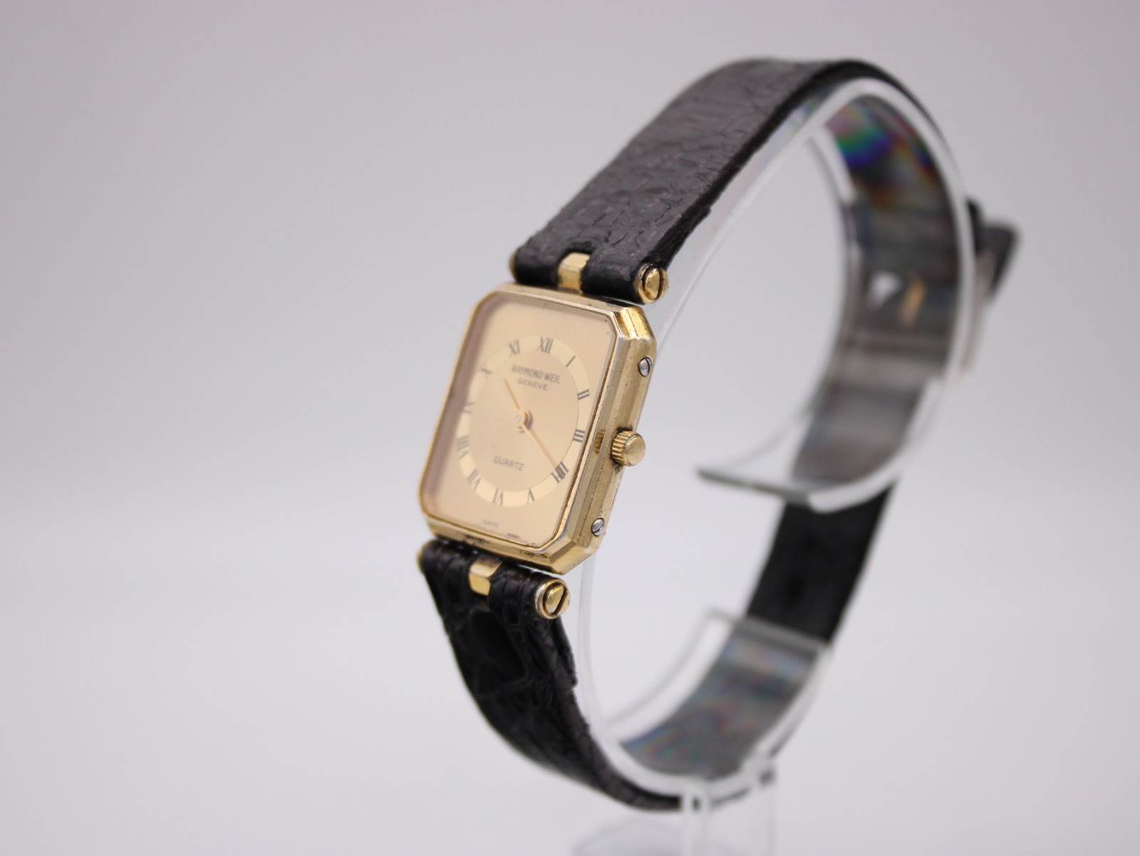 Angled side view of the Raymond Weil watch showing the gold case, crown, and black leather strap on a clear display stand.