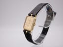 Angled side view of the Raymond Weil watch showing the gold case, crown, and black leather strap on a clear display stand.