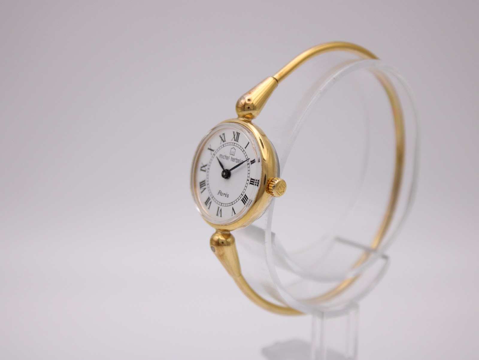 Three-quarter view of the gold Michel Herbelin manual wind watch and integrated bangle on a clear stand, showing the signed crown.