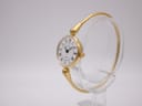 Three-quarter view of the gold Michel Herbelin manual wind watch and integrated bangle on a clear stand, showing the signed crown.