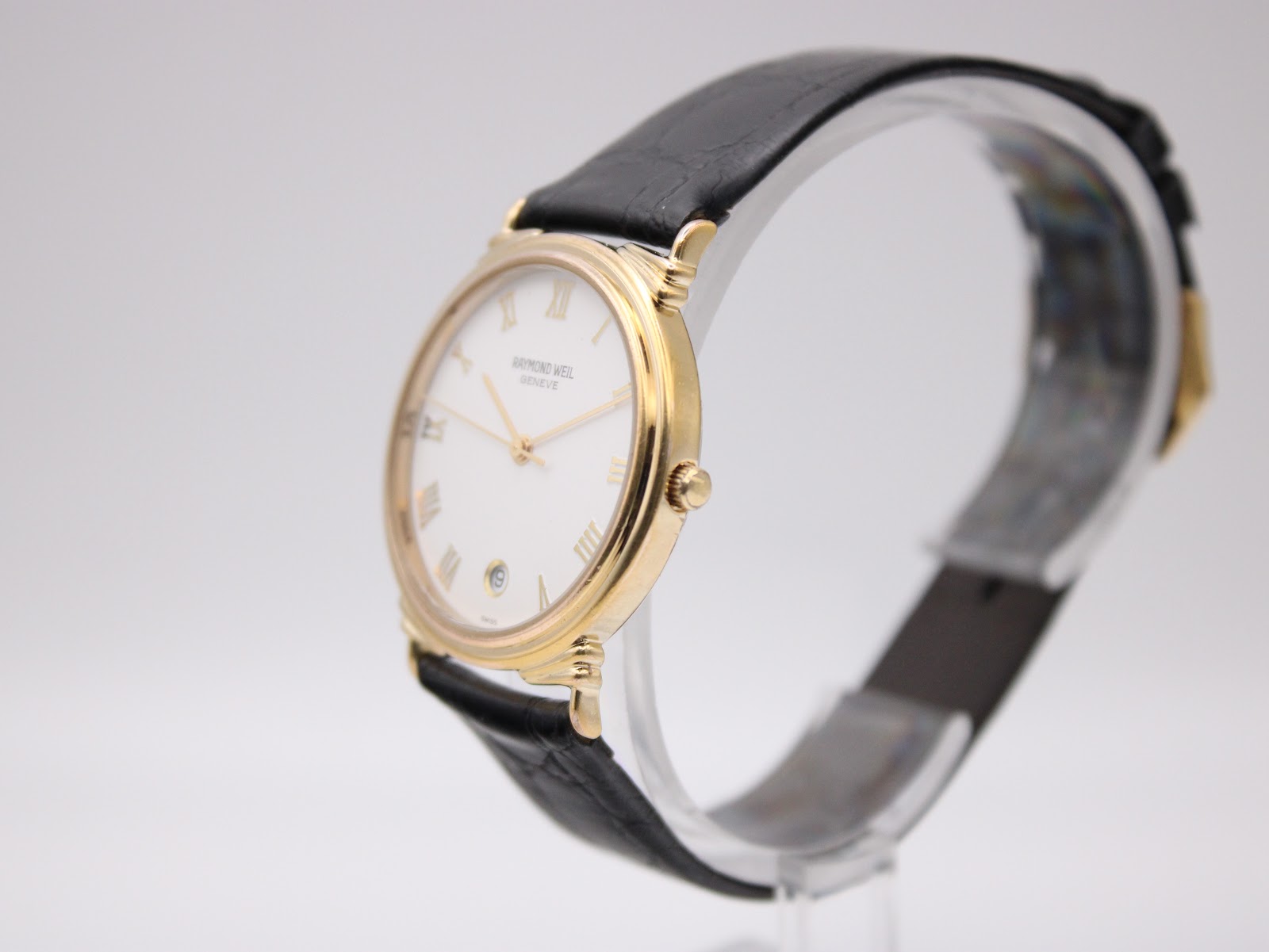 Angled side view of Raymond Weil watch on a display stand, showing the polished gold case and black strap.