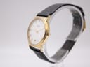 Angled side view of Raymond Weil watch on a display stand, showing the polished gold case and black strap.
