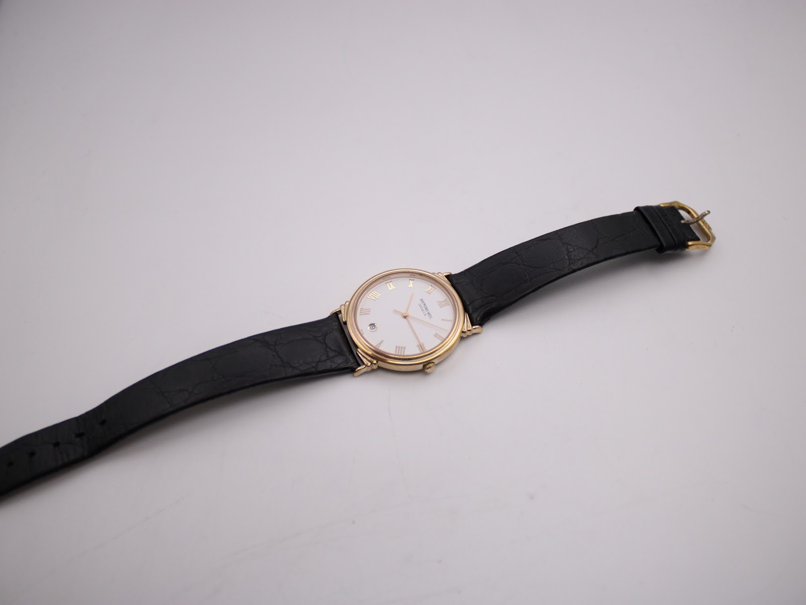 Full view of the Raymond Weil watch and black leather strap stretched out on a white surface.