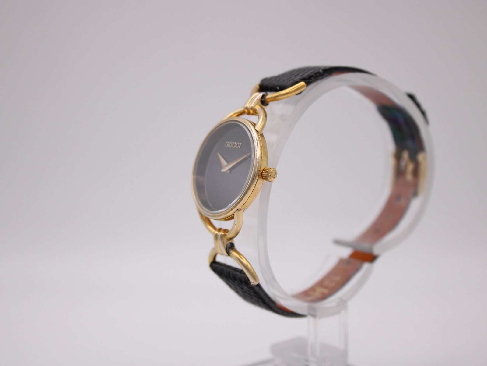 Side profile view of the Gucci watch on a stand, showing the round gold-tone case and crown.