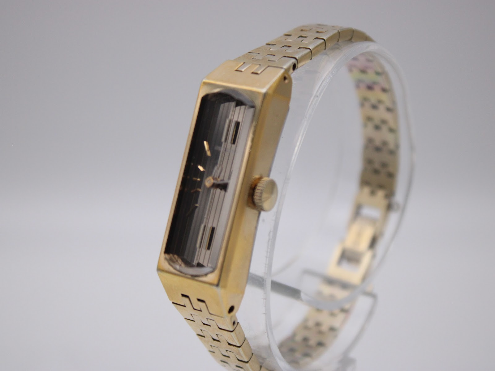 Side view of the gold-plated Seiko watch on a clear stand, highlighting the rectangular case and crown.