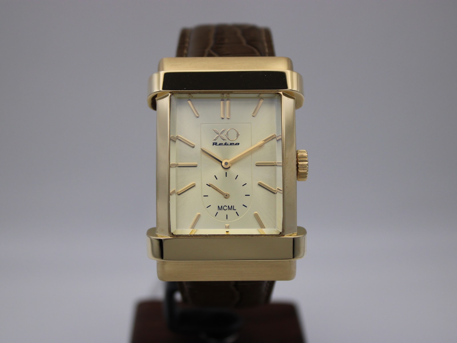 Front-on view of the XO RETRO watch on a display stand, with the cream dial, gold case, and brown strap clearly visible against a grey background.