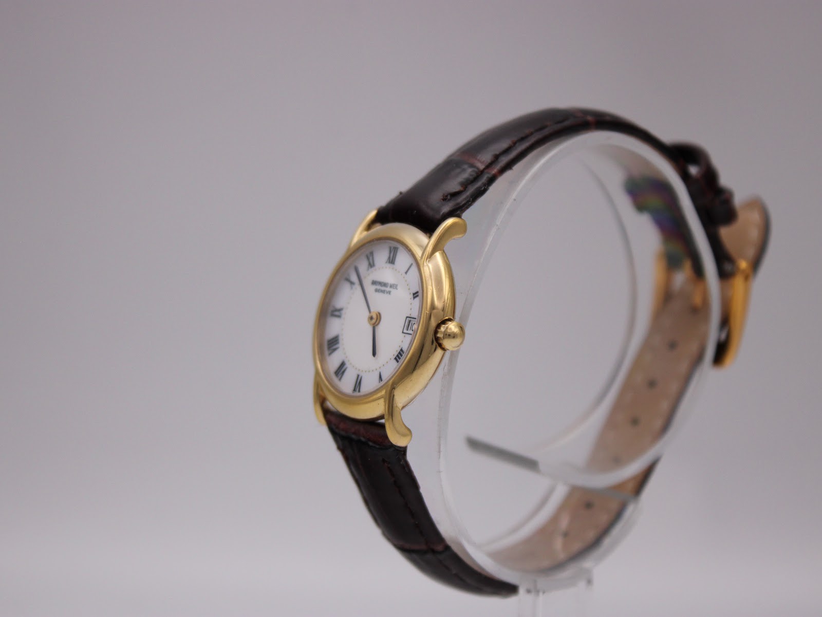 Angled side view of the Raymond Weil 9940 watch on a clear display stand, highlighting the gold case and strap.